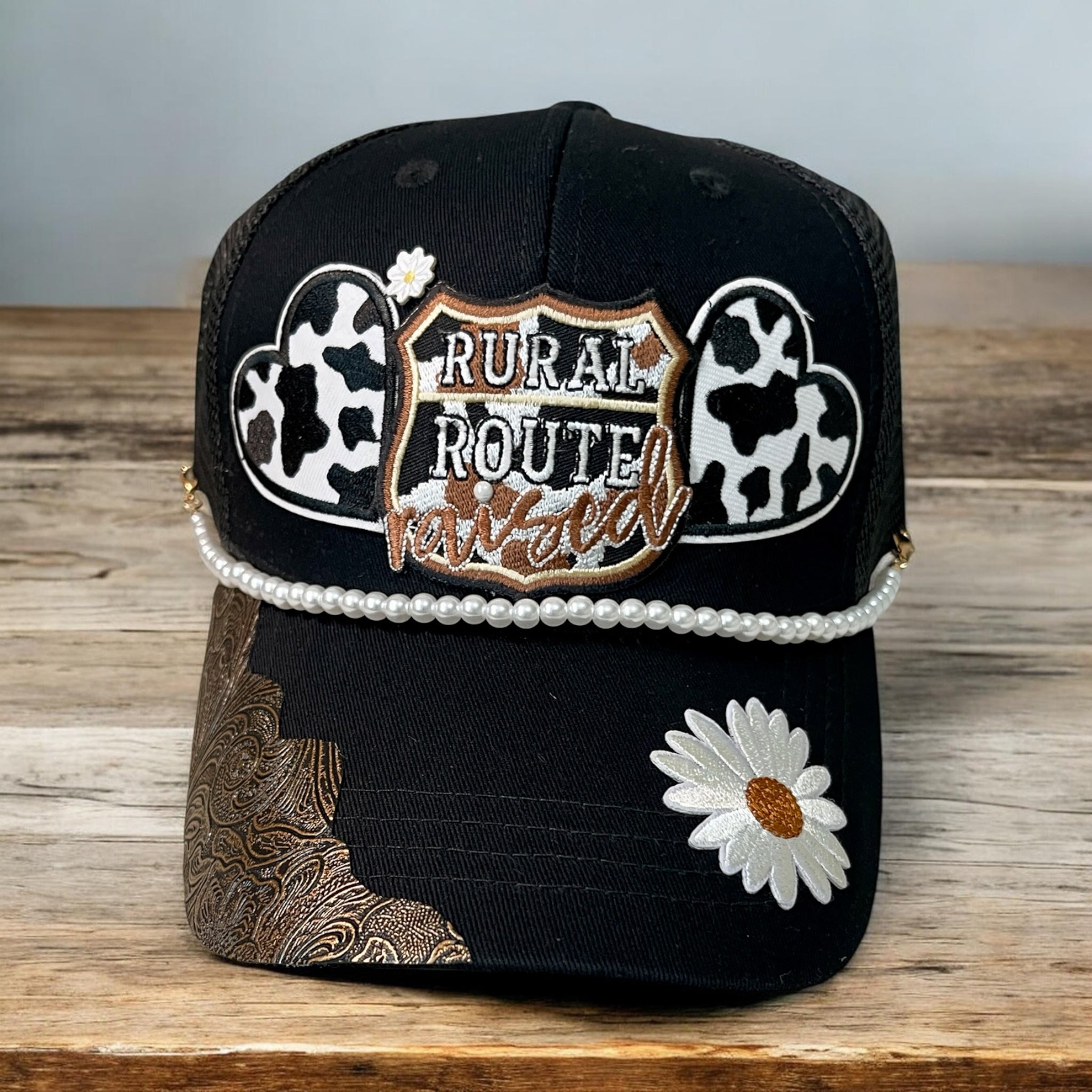 Rural Route Raised hat by Southern Brim Works - black cap with Rural Route Raised patch, cow print hearts, pearl trim, white daisy, and tooled leather corner
